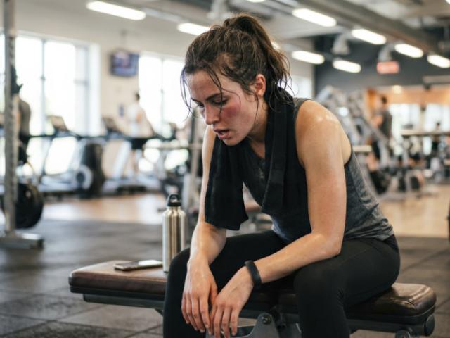 Mujer agotada sentada en el gimnasio, tras un entrenamiento intenso, evidenciando fatiga y posibles agujetas.  